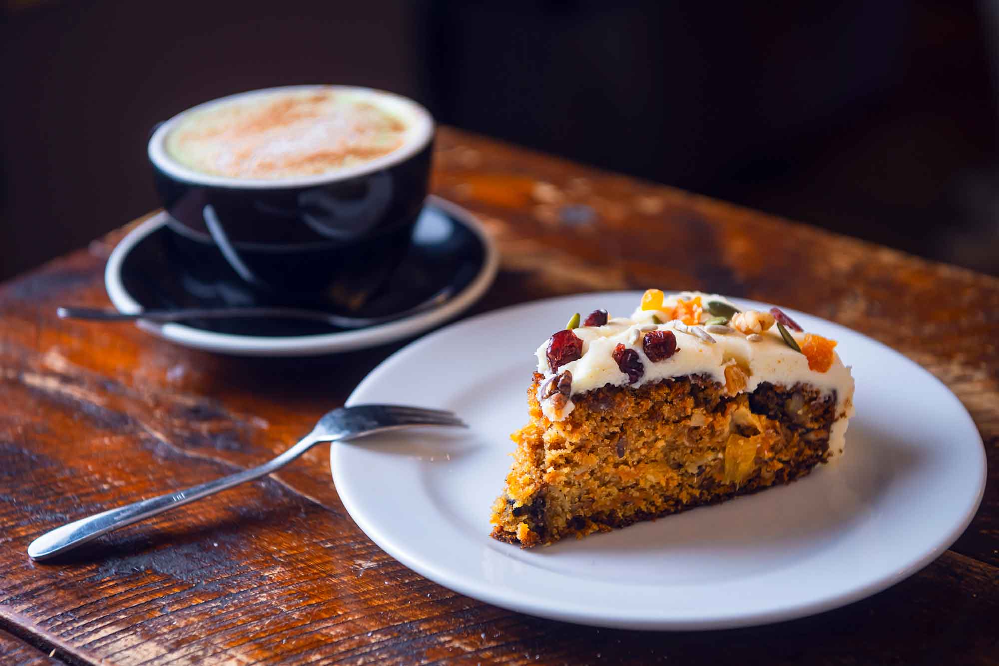 Slice of carrot cake with cream cheese frosting on a plate next to a cup of coffee .