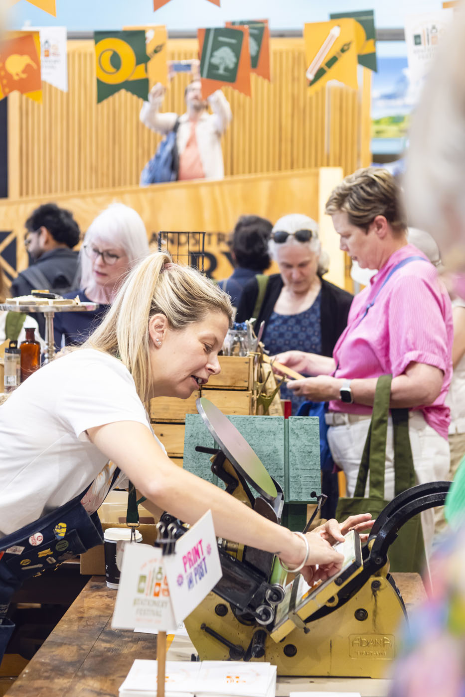 Lady using an old fashioned letterpress with people shopping in the background