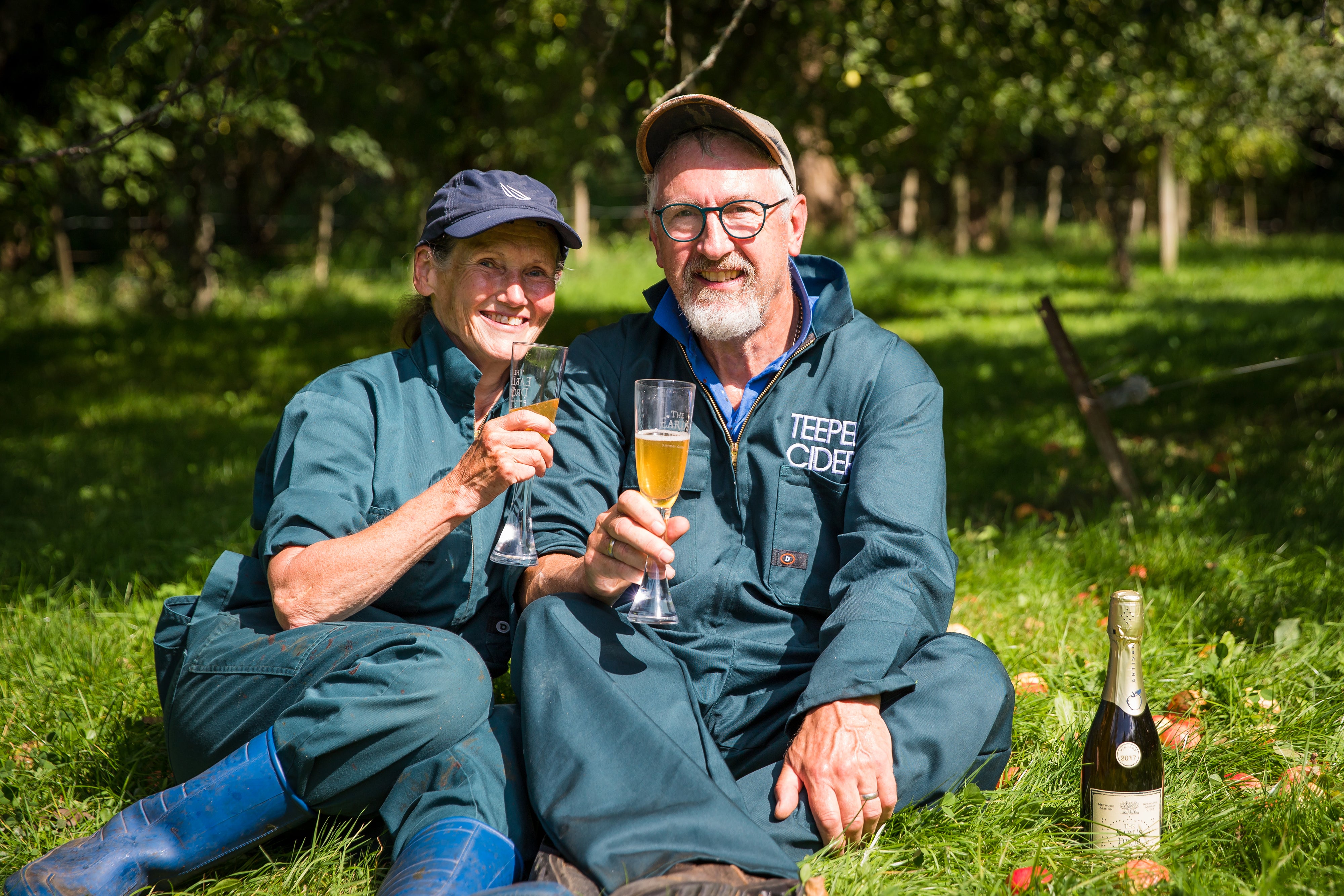 Owners sitting on grass drinking sparkling cider in orchard 