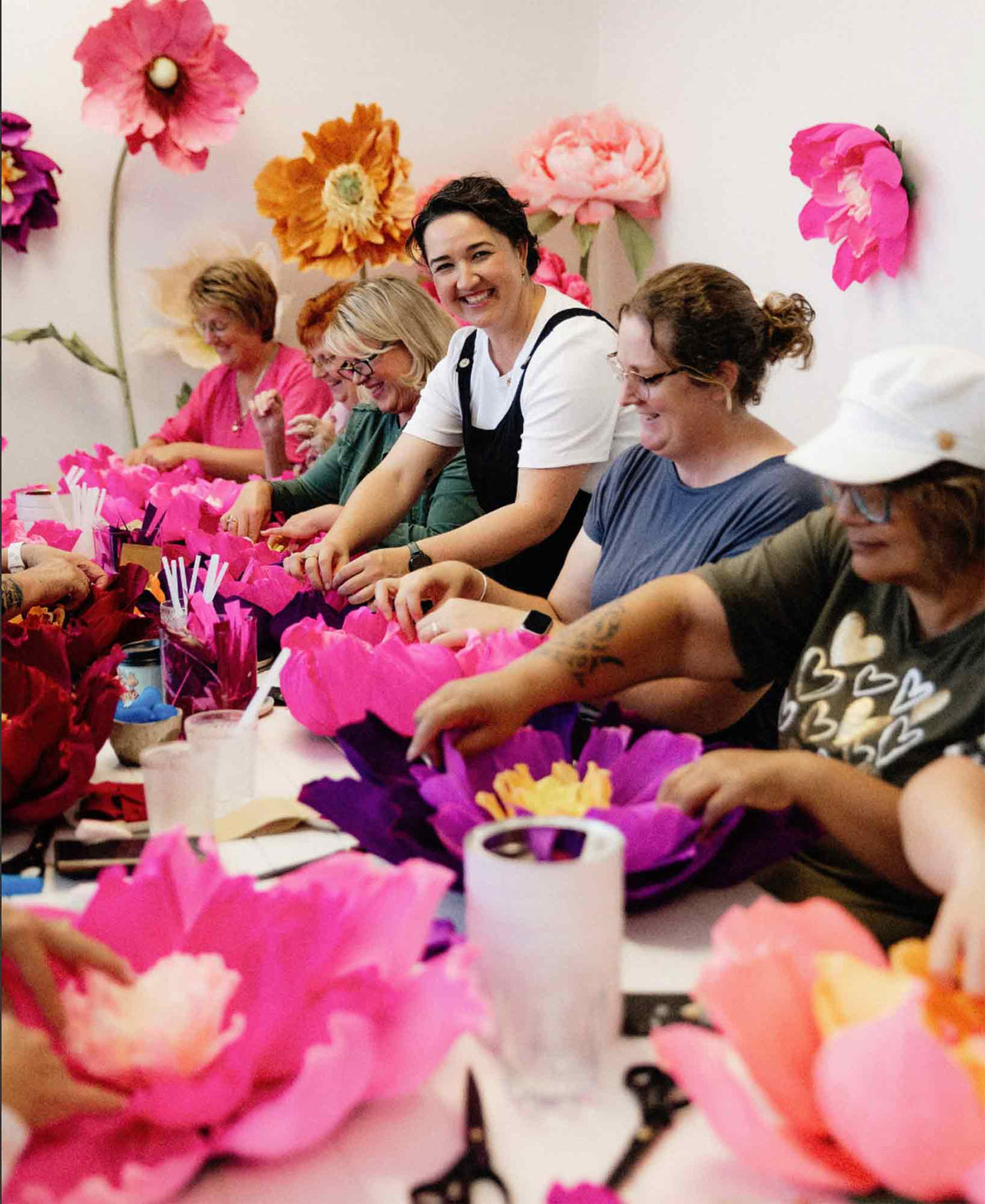 Several people sitting at a table making large paper flowers together in a bright indoor setting.